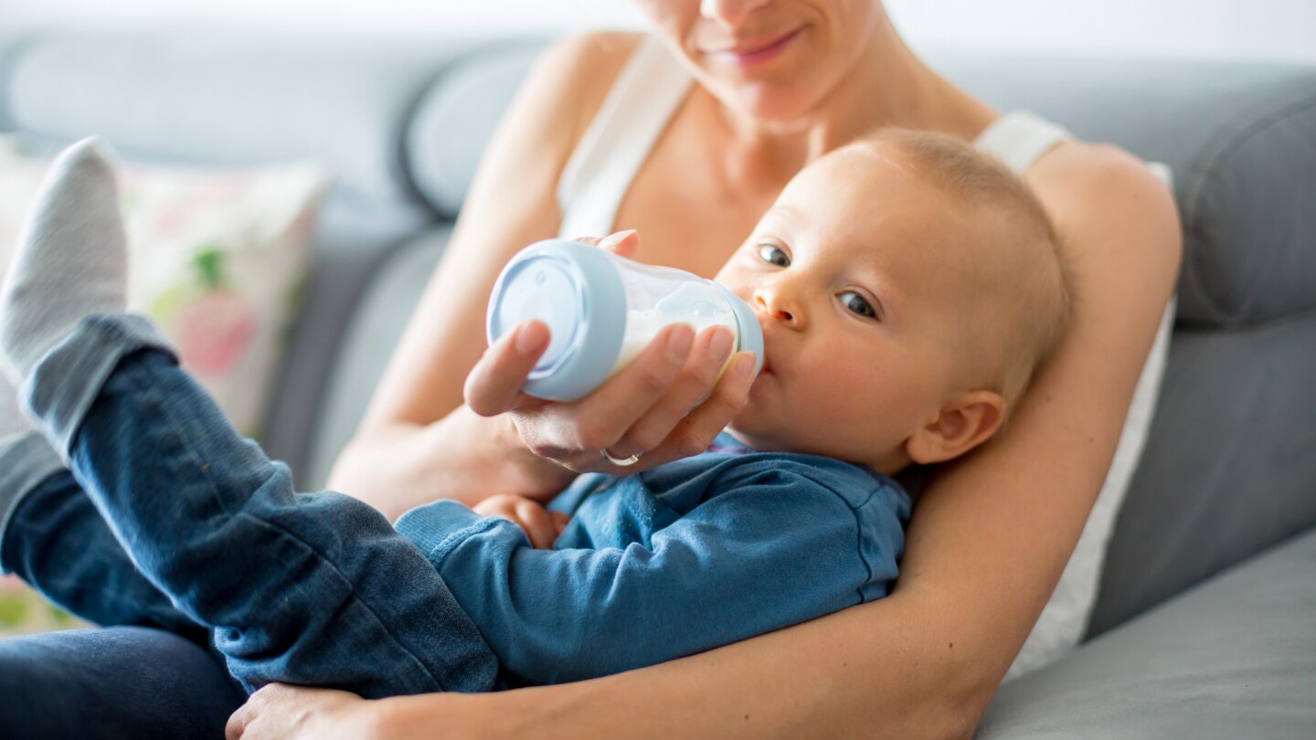 Mother, feeding her baby boy from bottle, sitting on the couch at home